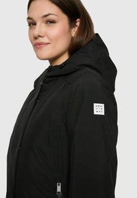 Frau, die eine schwarze Kapuzenjacke mit einem weißen rechteckigen Logo auf dem oberen Ärmel trägt, sichtbarem Reißverschluss und einem subtilen Lächeln.