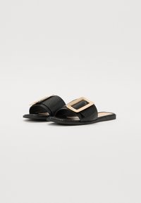 Sandales noires à glissière avec une tige texturée, arborant un grand accent de boucle métallique. Semelle plate avec un design minimaliste et bout ouvert.