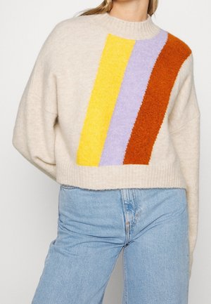 Femme portant un pull en tricot beige avec des rayures verticales jaunes, lavande et rouille, associé à un jean bleu clair, vu de la nuque aux hanches.