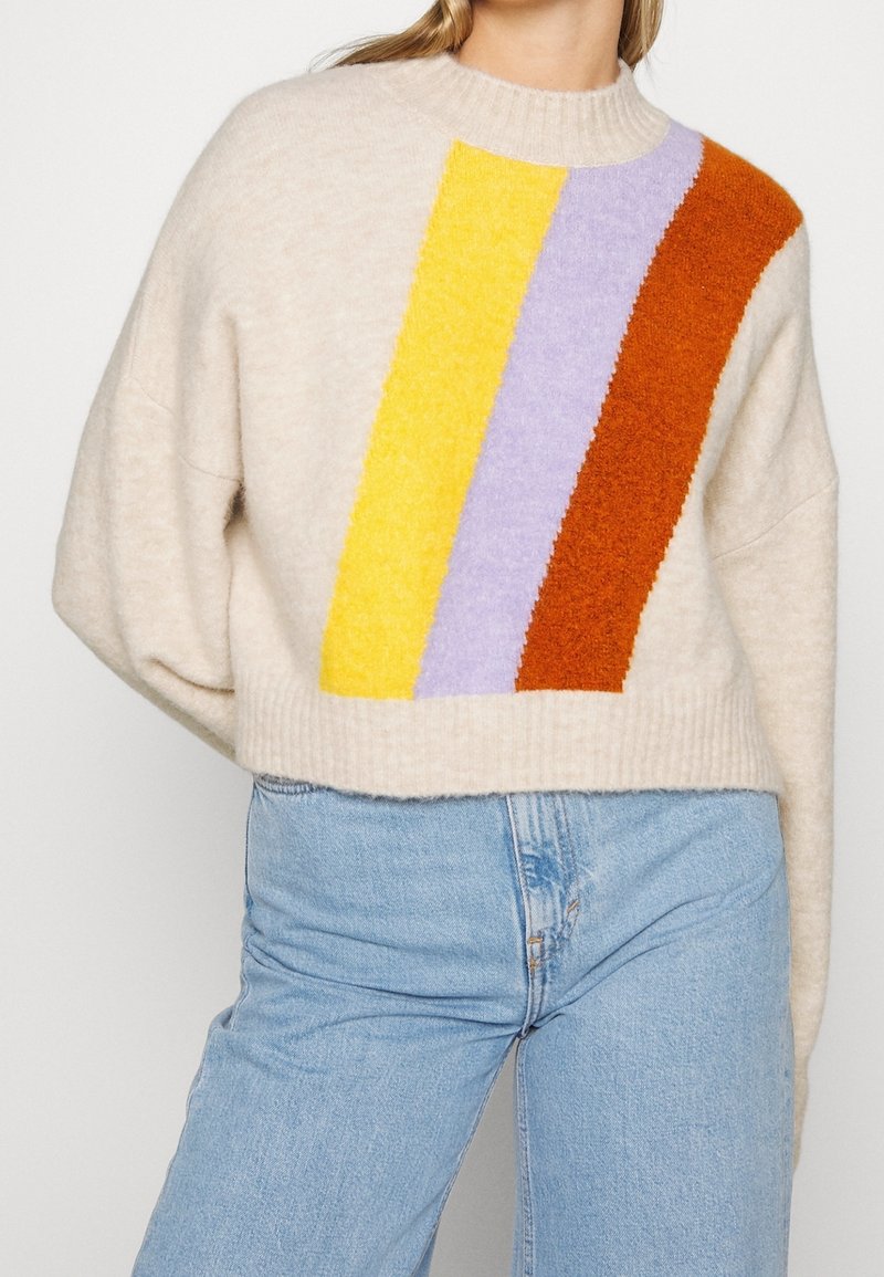 Femme portant un pull en tricot beige avec des rayures verticales jaunes, lavande et rouille, associé à un jean bleu clair, vu de la nuque aux hanches.