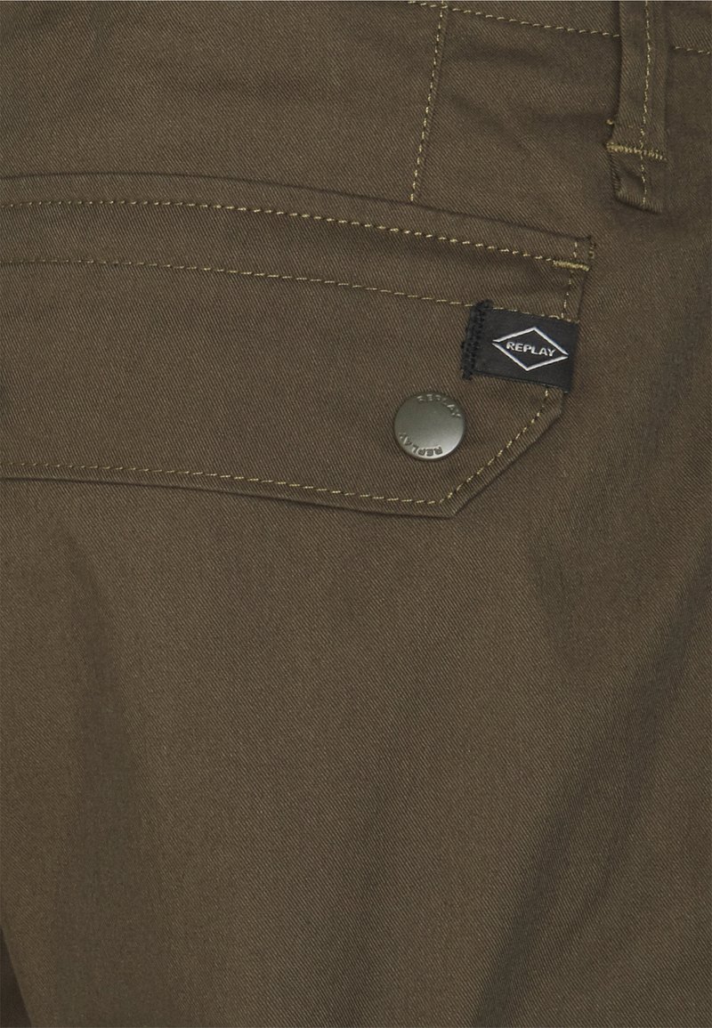 Brauner Stoff mit glatter Textur, der kontrastierende gelbe Nähte, einen Metallknopf und ein schwarzes Etikett mit der Aufschrift "REPLAY" aufweist.