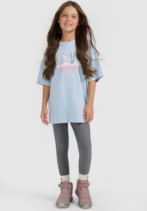 Młoda dziewczyna z długimi brązowymi włosami, ubrana w jasnoniebieski, oversize T-shirt z grafiką nart, szare legginsy, białe skarpetki i różowe śniegowce.