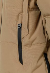 Beige pufferjacka med en slät yta, utrustad med en svart dragkedjeficka på sidan och en sydd sömdetalj längs tyget.