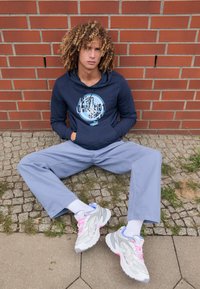 Felpa navy con design grafico, pantaloni larghi grigi chiari e sneakers grigie con dettagli rosa. L'ambientazione presenta ciottoli e erba.