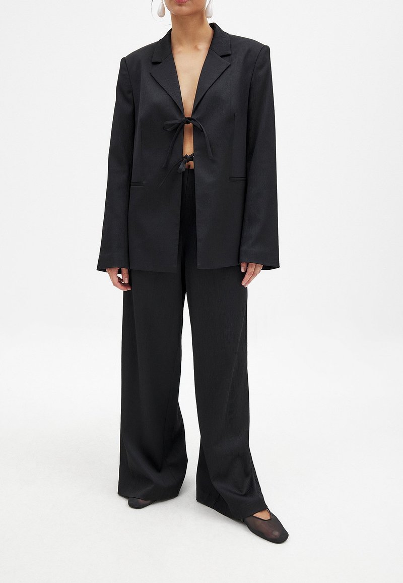 Črn blazer z dolgimi rokavi, sprednjim zavozom in obrnjenimi reverji. Pairs with širokimi črnimi hlačami in ravnimi črnimi čevlji.