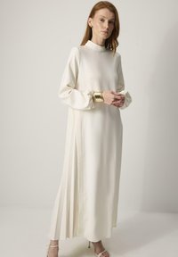 Lange, witte jurk met een hoge halslijn en geplooide zijpanelen. Soepele, satijnachtige textuur met lange mouwen en delicate gouden manchetten accent.