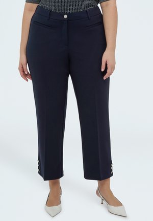 Žena nosí tmavě modré kalhoty délky ke kotníkům s knoflíkovými detaily u lemu a bílé lodičky na podpatku se zavazováním za patu a špičatou špičkou.