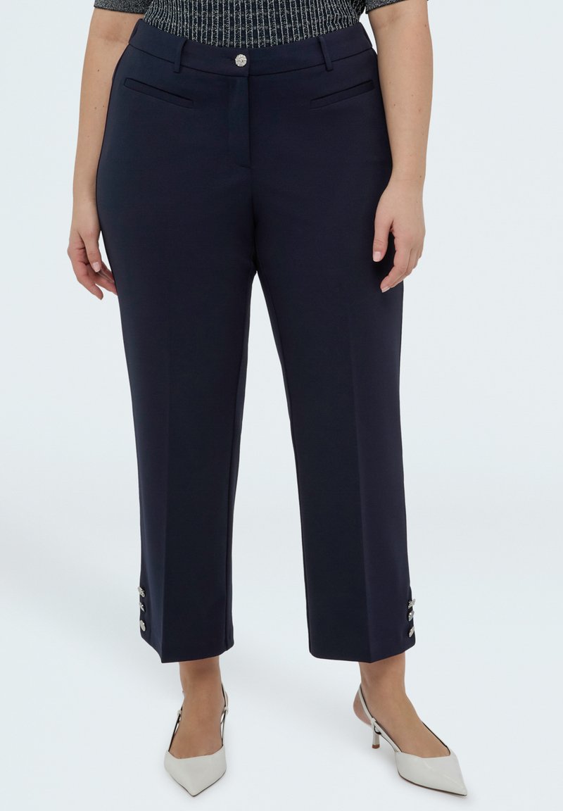 Femeie purtând pantaloni bleumarin până la gleznă, cu detalii cu nasturi lângă tivuri și pantofi albi cu toc ascuțit și barete slingback.