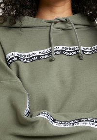 Olive green hoodie with drawstrings, featuring a white and black striped band with repeated "Adidas" text and logo across the chest.