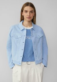 Femme portant une veste en denim bleu clair sur une chemise rayée bleu et blanc avec un pantalon blanc, se tenant devant un fond gris uni.