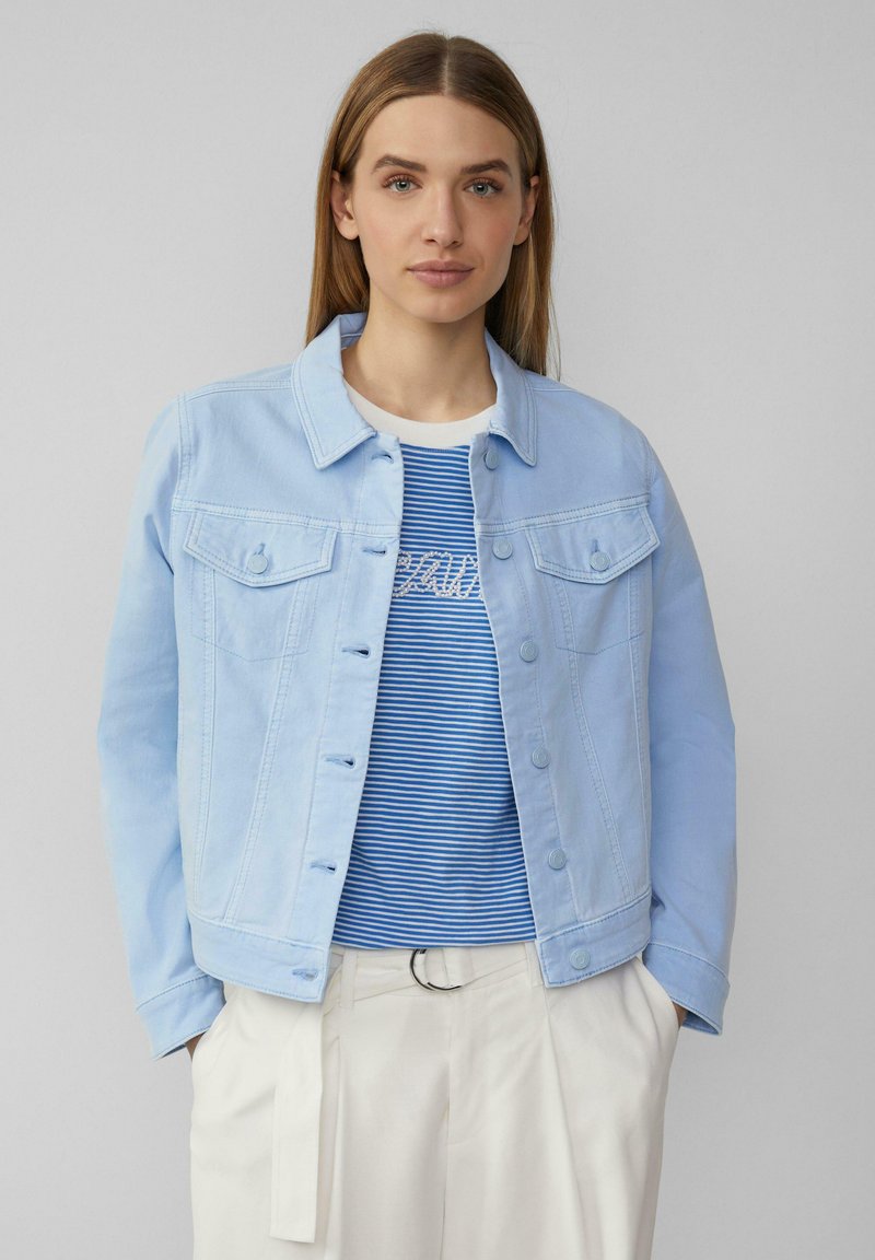 Femme portant une veste en denim bleu clair sur une chemise rayée bleu et blanc avec un pantalon blanc, se tenant devant un fond gris uni.