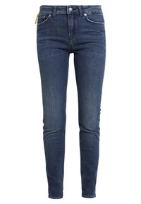 Jeans de mezclilla azul oscuro, corte ajustado, cintura media, diseño de cinco bolsillos, detalles de costura sutiles y una etiqueta amarilla en el bolsillo delantero.