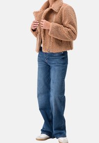 Persona che indossa una giacca sherpa color cammello, jeans a palazzo blu e sneakers bianche, mentre aggiusta la cerniera della giacca.