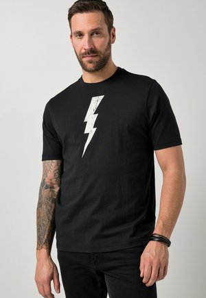 Mann mit kurzen Haaren und Bart, trägt ein schwarzes T-Shirt mit weißem Blitzmotiv, tätowierten linkem Arm und schwarzem Armband, steht vor einfarbigem Hintergrund.