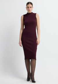 Bordowa, prążkowana sukienka bez rękawów z wysokim dekoltem, dopasowanym krojem i długością do kolan, połączona z czarnymi szpilkami.