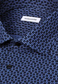 Gros plan sur un col de chemise à motif bleu et noir avec des boutons noirs et une étiquette blanche indiquant "seidensticker."