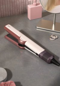 Dyson Haarglätter in Weiß und Roségold, mit einem eleganten Design, strukturiertem Griff und einem Kabel. Auf einer grauen Arbeitsplatte mit Zubehör platziert.