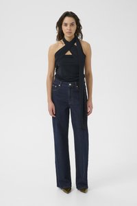 Melnā halterneck bluze ar šķērseniskām siksnām; augsta jostasvietas tumši zilu džinsu bikses ar piecām kabatām; eleganti, norādīti tumši zaļi papēži.