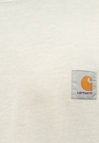 Svetlo béžový bavlnený tričko s námestíkovou nášivkou loga Carhartt v sivej a oranžovej farbe na ľavej strane hrudníka. Мäkká textura, dizajn s kruhovým výstrihom.
