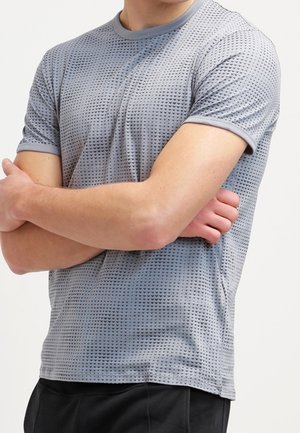 Persoon draagt een lichtgrijs, kort mouw, patroon overhemd met gekruiste armen, gecombineerd met een zwarte broek tegen een egaal achtergrond.