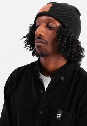 Black ribbed knit beanie with a square leather patch. Model wears a black corduroy shirt with white undershirt and a black logo on the pocket.