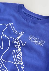 T-shirt bleu avec un dessin en ligne blanche de mains tenant un trophée et le texte « GAME ON 24/SEVEN PRO. FINALS » sur la poitrine.