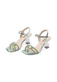 Sandali con tacco verde metallizzato, punta quadrata, tacco a blocco trasparente e cinturini con texture annodata. Presentano una chiusura con fibbia alla caviglia.