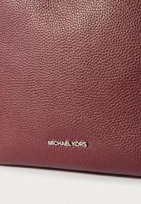 Burgundivörös, textúrázott bőr kézitáska sima felülettel és arany "MICHAEL KORS" logóval a sarokban.