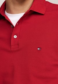 Gros plan sur une personne portant un polo rouge avec des boutons blancs et un petit logo rectangulaire sur la poitrine. Sous-vêtement blanc visible.