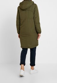 Person klädd i en olivgrön huva lång kappa, marinblå uppvikta byxor och vita sneakers, stående mot en enfärgad vit bakgrund.