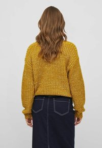 Senfgelbes Strickpullover in Übergröße mit langen Ärmeln und strukturiertem Muster. Kombiniert mit einem dunklen Denim-Rock.