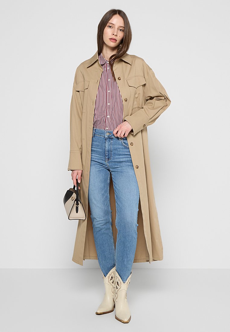 Beżowy długi płaszcz na czerwono-białą w paski koszulę, niebieskie dżinsy i białe kowbojskie buty. Mała czarno-biała torebka trzymana w jednej ręce.