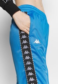 Blå satin träningsbyxor med en svart sidostrand som har upprepade logotypmönster. Elastisk midja och sidoficka synlig. Vit logotyp fram.