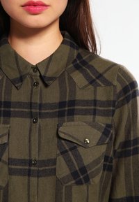 Olivgrön flanellskjorta med marinblå rutmönster, knapp-up framtill, spetsig krage och två bröstfickor med metallknappar.