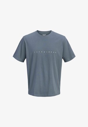Chłopiec stoi, ubrany w niebieski T-shirt z napisem, szerokie wyblakłe jeansy i białe trampki na jasnym, jednolitym tle.