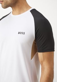 Pánské sportovní tričko s bílým tělem, černými rukávy a hnědým bočním panelem. Obsahuje kulatý výstřih a logo "BOSS" na hrudi.