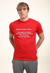 Rød bomulds T-shirt med hvid tekst, der siger "De fire livsfaser..." efterfulgt af humoristiske sætninger relateret til Santa. Korte ærmer.