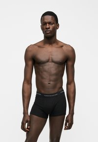 Uomo senza maglietta con pelle scura che indossa boxer neri Tommy Hilfiger in piedi davanti a uno sfondo bianco semplice.