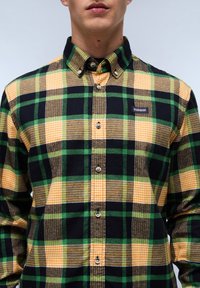 Kraagloze flanellen shirt met een geruite patroon in groen, geel, bruin en zwart, met knoopsluitingen en een klein merklabel op de borst.