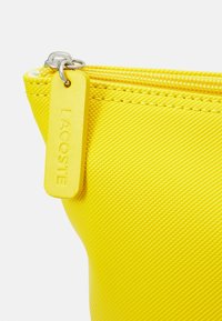 Pochette jaune texturée avec fermeture éclair. Présente une étiquette en caoutchouc avec le nom de la marque "LACOSTE" en relief. Tirette de fermeture éclair en métal lisse.