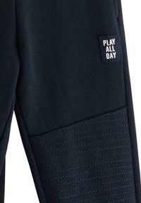Mörkblå träningsbyxor med en strukturerad nederdel och en svart etikett med "PLAY ALL DAY" i vit text. Slät tyg överlag.