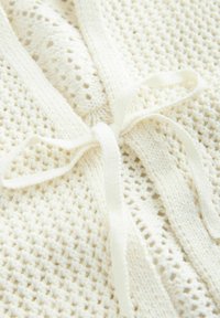 Tessuto lavorato a maglia color crema con motivo a trama aperta e texture, impreziosito da un dettaglio di fiocco annodato al centro.