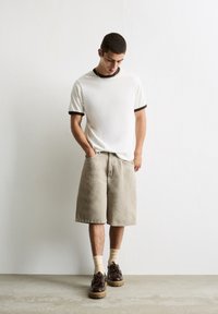 Chemise blanche à manches courtes avec col côtelé marron, shorts beiges, chaussures marron et chaussettes crème, se tenant devant un fond texturé clair.