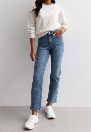 Personne portant un jean bleu taille haute, des baskets blanches et un sweat-shirt à manches longues couleur crème, adossée à un mur neutre.