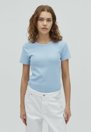 Young woman with curly brown hair wearing a light blue short-sleeve shirt and white pants, standing against a plain background.