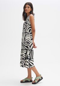 Black and white patterned sleeveless dress with a flowing shape and tiered hem, paired with green and black cross-strap sandals.