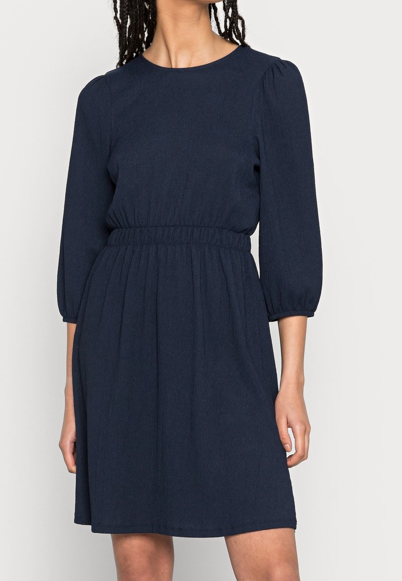 Femme portant une robe bleu marine longueur genoux avec des manches trois-quarts bouffantes et une taille cintrée élastique.