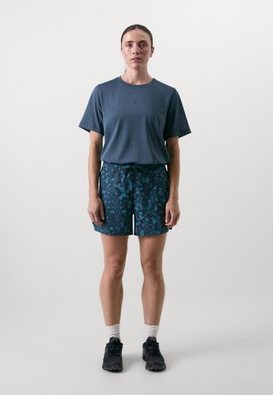 T-Shirt mit kurzen Ärmeln in Teal mit Logo auf der Brust, kombiniert mit blauen gemusterten Shorts. Das Model trägt schwarze Schuhe und weiße Knöchel-Socken.