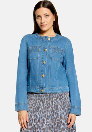 Woman wearing a blue denim jacket with gold buttons and a pleated leopard print skirt against a plain background.