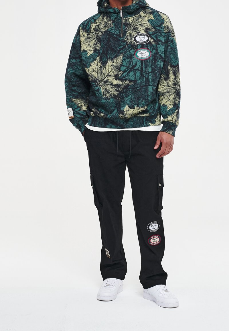 Sudadera con capucha y cremallera con un patrón de hojas verdes y beige, combinada con pantalones cargo negros con parches y zapatillas blancas.